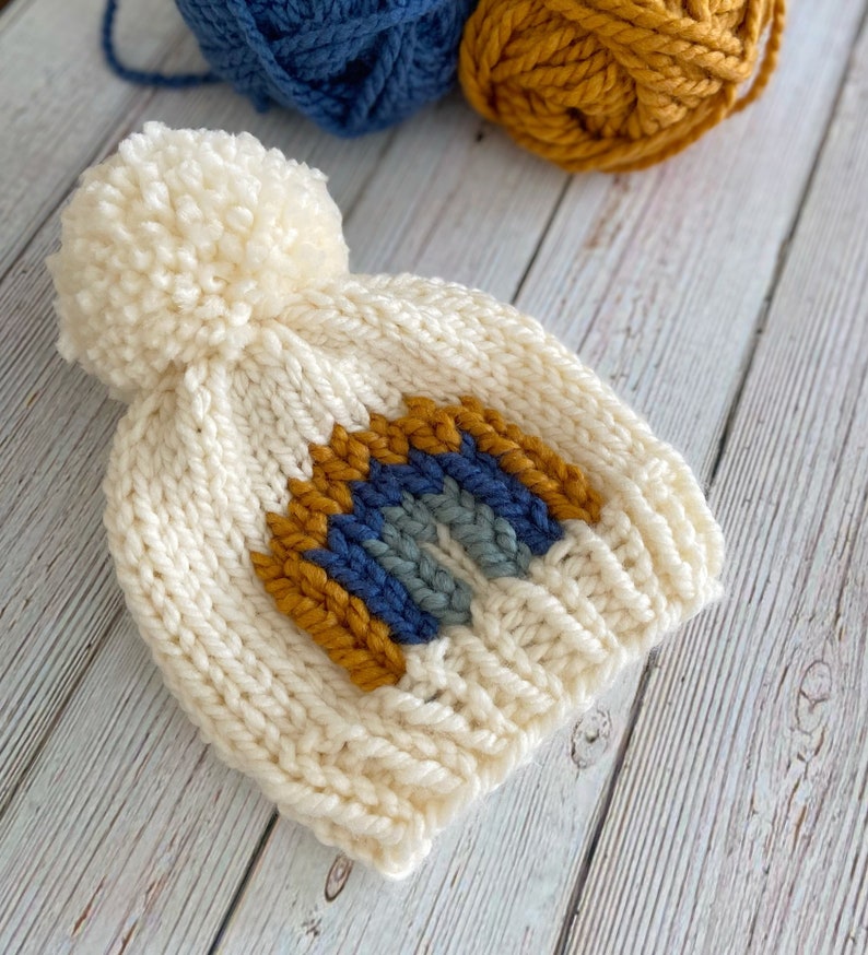 Op de afbeelding: Witte gebreide muts met een pompon bovenop en een regenboogontwerp in blauw, grijs en geel.