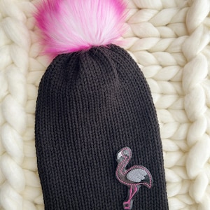 May include: Black knit beanie with a large pink and white faux fur pom-pom. The hat features a flamingo patch with pink and gray accents. The beanie is displayed on a white, chunky knit blanket.