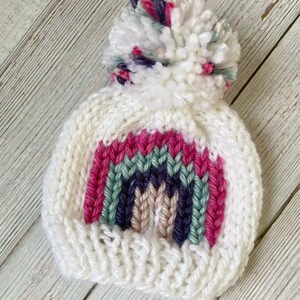 Op de afbeelding: Witte gebreide muts met een regenboogontwerp en een pluche witte pompon bovenop. De regenboog is gemaakt van roze, groen, paars en beige garen.