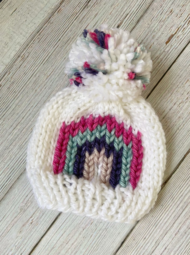 Op de afbeelding: Witte gebreide muts met een regenboogontwerp en een pluche witte pompon bovenop. De regenboog is gemaakt van roze, groen, paars en beige garen.