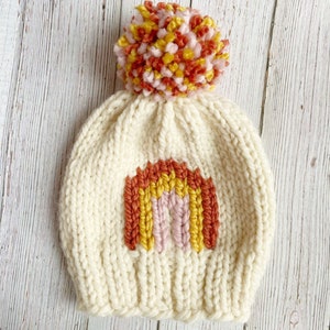 Op de afbeelding: Een witte gebreide muts met een regenboogontwerp en een roze, gele en oranje pompon.