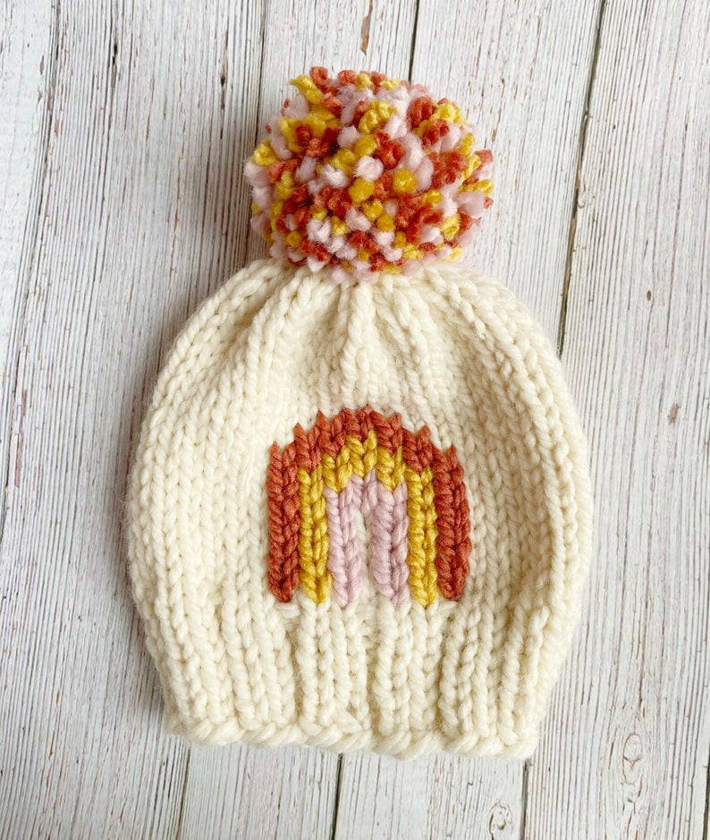 Op de afbeelding: Een witte gebreide muts met een regenboogontwerp en een roze, gele en oranje pompon.