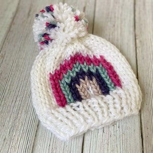 Op de afbeelding: Witte gebreide muts met een regenboogstreepdessin en een pluche witte pompon bovenop. De regenboogstreep is gemaakt van roze, groen, paars en beige garen.