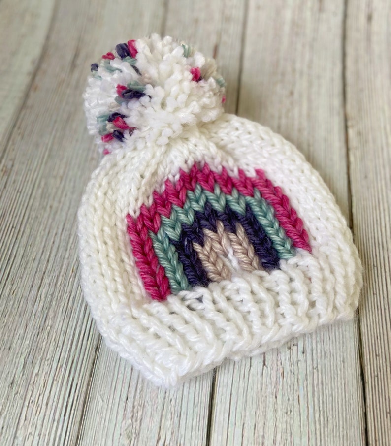 Op de afbeelding: Witte gebreide muts met een regenboogstreepdessin en een pluche witte pompon bovenop. De regenboogstreep is gemaakt van roze, groen, paars en beige garen.