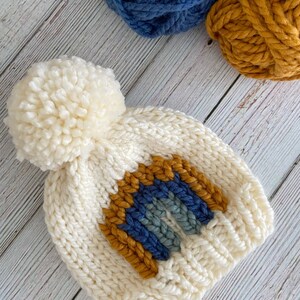 Op de afbeelding: Witte gebreide muts met een pompon bovenop en een regenboogontwerp in blauw, grijs en mosterdgeel.
