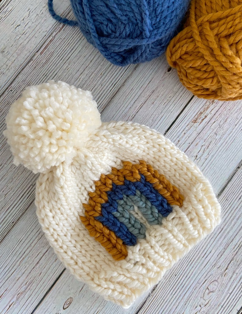 Op de afbeelding: Witte gebreide muts met een pompon bovenop en een regenboogontwerp in blauw, grijs en mosterdgeel.