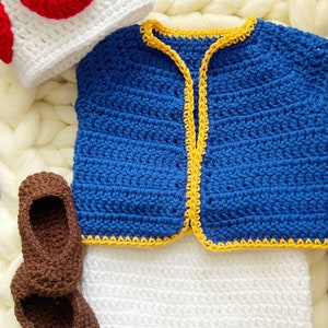 May include: A crocheted baby outfit in blue, white, and brown. The outfit includes a hat, a vest, shorts, and booties. The hat is shaped like a mushroom with red spots.