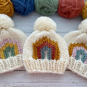 Op de afbeelding: Drie witte gebreide mutsen met regenboogontwerpen. Elke muts heeft een witte pompon bovenop. De regenboogontwerpen zijn gemaakt met verschillende kleuren garen, waaronder roze, geel, groen, blauw en rood.