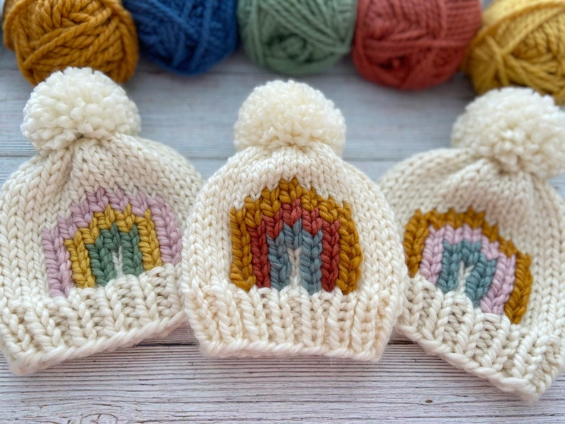Op de afbeelding: Drie witte gebreide mutsen met regenboogontwerpen. Elke muts heeft een witte pompon bovenop. De regenboogontwerpen zijn gemaakt met verschillende kleuren garen, waaronder roze, geel, groen, blauw en rood.