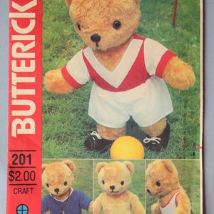May include: A Butterick 201 craft pattern envelope featuring a plush teddy bear wearing a red and white sports jersey and shorts with a yellow ball. Additional images show the bear in a blue outfit, a plain bear, and a white tank top with brown shorts.