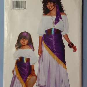 May include: Butterick sewing pattern 4653 featuring an off-the-shoulder white blouse, a long light purple skirt, a purple sash with gold fringe, and a matching headscarf. The pattern includes instructions for both adult and child sizes.