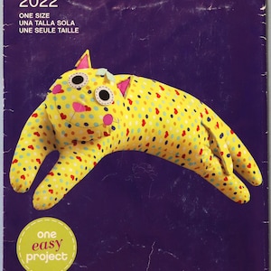 May include: A Simplicity sewing pattern for a cat-shaped stuffed animal. The pattern cover is purple and features a yellow cat with colorful polka dots. The text "2022" and "One Size" are also visible. The words "Sew Simple" and "one easy project" are also visible.
