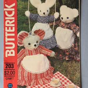 May include: A Butterick 203 craft pattern envelope featuring three plush animal figures dressed in floral and striped dresses with white aprons. The scene includes a red and white checkered picnic cloth with a teapot and three teacups.