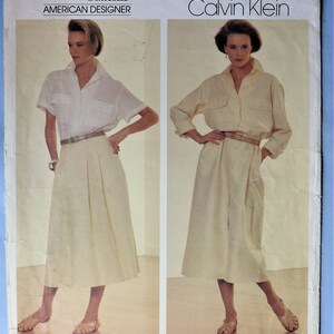 May include: A Vogue Patterns American Designer Calvin Klein sewing pattern envelope 1320. The front displays two views of a garment: a white short-sleeved collared shirt with a pleated skirt, and a long-sleeved collared shirt dress with a belt.
