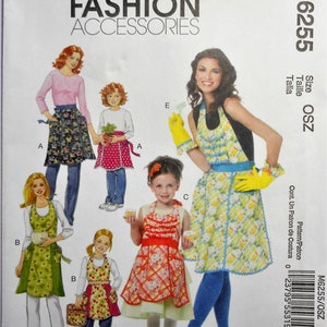 May include: McCall's Fashion Accessories sewing pattern M6255. The pattern features various apron designs, including floral, polka dot, and cupcake prints. The cover showcases multiple apron styles for adults and children, with the text "Fashion Accessories".