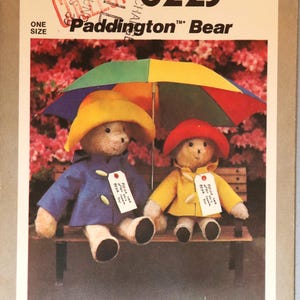 May include: Simplicity 8223 sewing pattern envelope for a Paddington Bear stuffed toy. The cover features two plush bears sitting on a bench under a multi-colored umbrella. One bear wears a blue coat and yellow hat, the other a yellow coat and red hat.