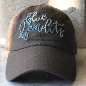 Puede incluir: Gorra de béisbol marrón con el texto bordado "blue bandits" en hilo azul claro y blanco. La gorra tiene una visera curva y un diseño clásico de seis paneles, adecuada para uso casual.