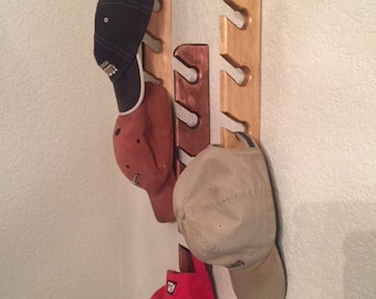May include: A wooden hat rack with six slots for storing baseball caps. The rack is mounted on a wall and has four hats hanging on it, including a red, black, brown, and beige hat.