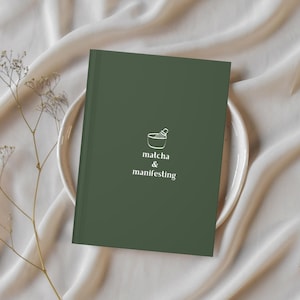 May include: A dark green journal with the words "matcha & manifesting" and a small graphic of a bowl and whisk. The journal is on a white plate, with a cream-coloured fabric in the background. A sprig of dried flowers is on the left.