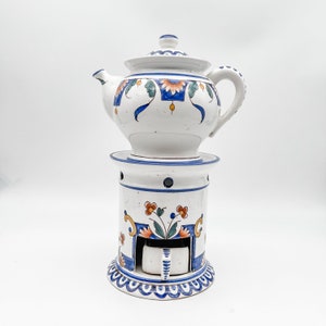 May include: White ceramic teapot with a matching warmer. The teapot and warmer are decorated with blue, orange, and green floral patterns. The teapot has a lid and a curved handle. The warmer has a circular base and a small opening for a candle.