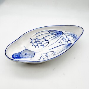 May include: A white ceramic serving dish with a blue and white painted design of a duck. The duck is facing left and has its wings spread out. The dish is oval shaped and has a slight curve to it.