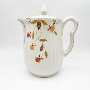 May include: White ceramic teapot with a gold trim and a floral pattern of brown leaves and orange flowers. The teapot has a lid with a knob on top.