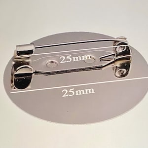 May include: A silver round pin back with a safety pin attached. The pin back measures 25 mm in diameter.