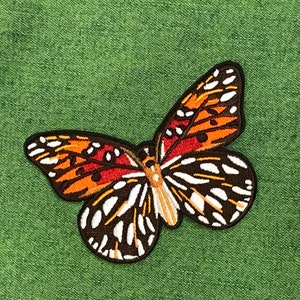 Puede incluir: Una mariposa marrón y naranja con marcas blancas y negras en sus alas. La mariposa está bordada en una tela verde.