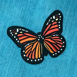 May include: A black and orange butterfly patch with white dots. The butterfly is embroidered on a blue denim fabric.