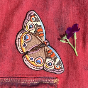 Könnte beinhalten: Ein gestickter Schmetterlingspatch in Braun und Rosa mit blauen und orangen Akzenten. Der Patch befindet sich auf einem roten Stoffhintergrund.