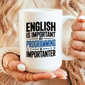 May include: A white ceramic mug featuring black and blue text that reads: ENGLISH IS IMPORTANT BUT PROGRAMMING IS IMPORTANTER, with the word PROGRAMMING in blue and decorative brackets surrounding the word IS in the center of the design.