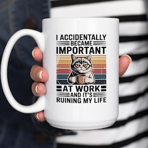 K&ouml;nnte beinhalten: Wei&szlig;e Keramik-Tasse mit schwarzem Henkel. Die Tasse zeigt eine Katze, die eine Kaffeetasse h&auml;lt, und den Text "I ACCIDENTALLY BECAME IMPORTANT AT WORK AND IT'S RUINING MY LIFE" in Schwarz.