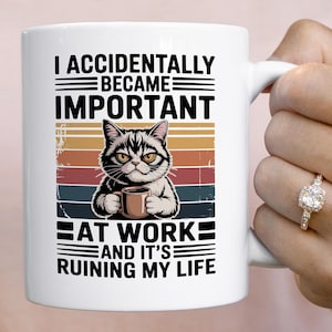 K&ouml;nnte beinhalten: Wei&szlig;e Keramiktasse mit einer Illustration einer grummeligen Katze, die eine Kaffeetasse h&auml;lt. Die Tasse tr&auml;gt den schwarzen Text "I ACCIDENTALLY BECAME IMPORTANT AT WORK AND IT'S RUINING MY LIFE" mit einem Retro-Farbblock-Design.