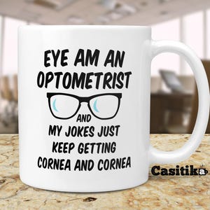 May include: White ceramic coffee mug with a black and white design. The mug reads "Eye am an optometrist and my jokes just keep getting cornea and cornea".