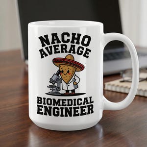 May include: A white ceramic mug featuring a cartoon tortilla chip wearing a sombrero and a white lab coat, looking through a microscope. The text NACHO AVERAGE BIOMEDICAL ENGINEER is printed in bold black letters above and below the illustration.