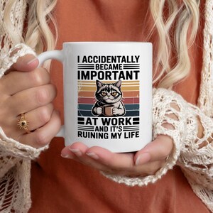 K&ouml;nnte beinhalten: Wei&szlig;e Keramik-Tasse mit einer Katzenillustration, die eine Kaffeetasse h&auml;lt, mit dem Text: &bdquo;I ACCIDENTALLY BECAME IMPORTANT AT WORK AND IT'S RUINING MY LIFE&ldquo;. Das Design beinhaltet einen Retro-Sonnenuntergangshintergrund.