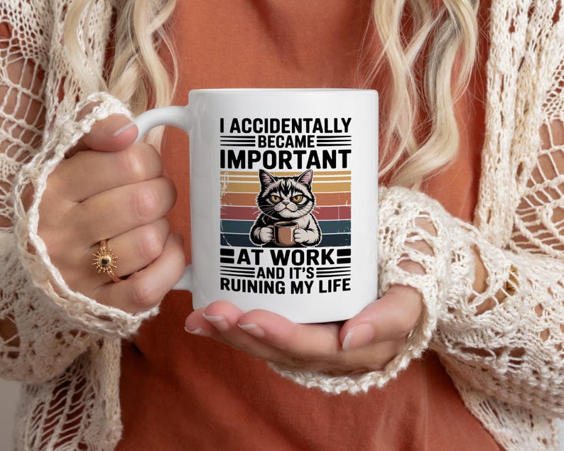 K&ouml;nnte beinhalten: Wei&szlig;e Keramik-Tasse mit einer Katzenillustration, die eine Kaffeetasse h&auml;lt, mit dem Text: &bdquo;I ACCIDENTALLY BECAME IMPORTANT AT WORK AND IT'S RUINING MY LIFE&ldquo;. Das Design beinhaltet einen Retro-Sonnenuntergangshintergrund.