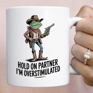May include: White ceramic mug featuring a cartoon frog dressed as a cowboy, holding a gun. The frog wears a brown hat, jacket, and boots, with a red bandana. Text on the mug reads: "HOLD ON PARTNER I'M OVERSTIMULATED."