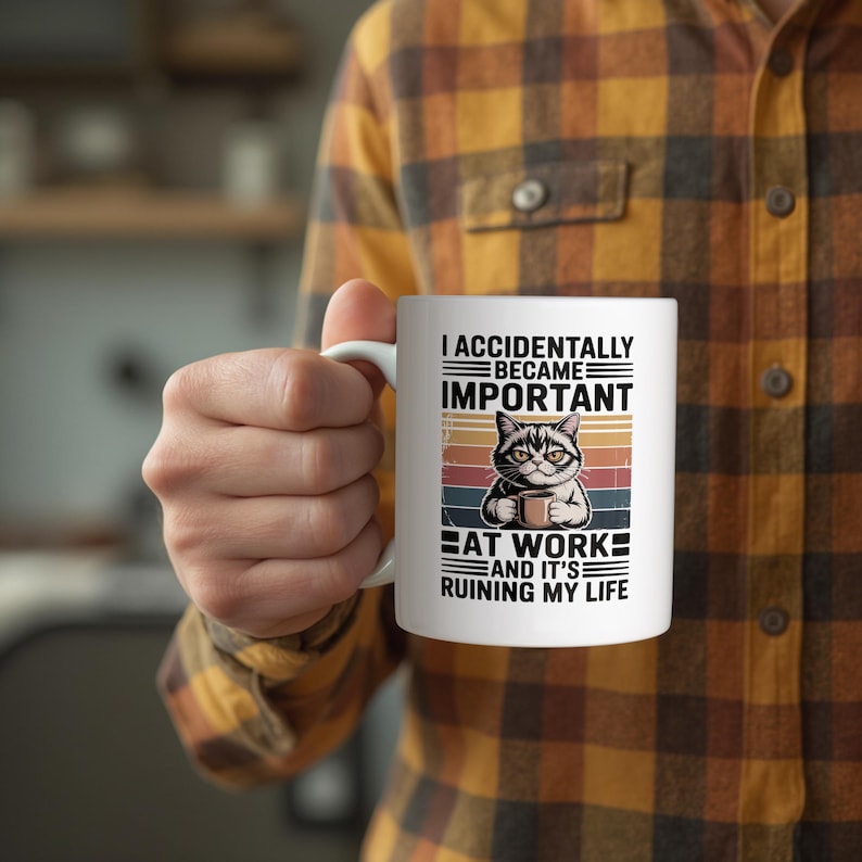 K&ouml;nnte beinhalten: Wei&szlig;e Keramik-Tasse mit einer Katzenillustration, die eine Kaffeetasse h&auml;lt, mit dem Text "I ACCIDENTALLY BECAME IMPORTANT AT WORK AND IT'S RUINING MY LIFE." Die Tasse hat einen wei&szlig;en Griff.