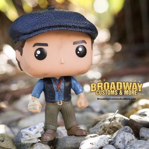 May include: A Funko Pop figure of a character wearing a blue newsboy cap, blue shirt, black vest, and brown pants. The figure is standing on rocks with a stack of newspapers. The Broadway Customs & More logo is visible.