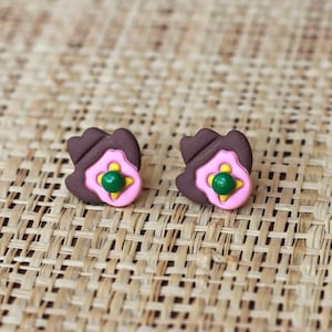 Puede incluir: Un par de pendientes de color marrón y rosa en forma de flor con un centro verde. Los pendientes están sobre un fondo marrón tejido.
