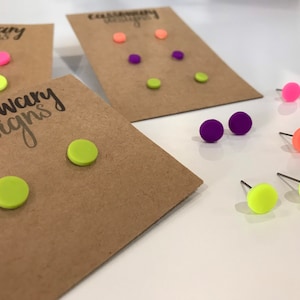 May include: Assowary Designs logo on a brown card with two pairs of neon green round stud earrings. The earrings are displayed on a white surface.