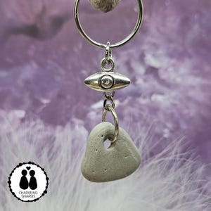 May include: A silver-toned keychain featuring a gray stone pendant with a hole, a silver eye-shaped charm, and a circular ring. The keychain has a purple background with the "Charming Shards" logo in the lower left corner.