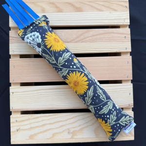 May include: A cylindrical fabric toy featuring a dark gray background with a yellow dandelion and white seed head pattern. The top end has four bright blue ribbon strips extending outward, and a small white fabric tag is attached to the bottom edge.