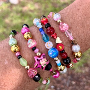 May include: A collection of beaded bracelets with a variety of colorful beads. The bracelets feature gold, pink, black, and blue beads, including heart and star-shaped charms. The bracelets are on display against a blurred green background.