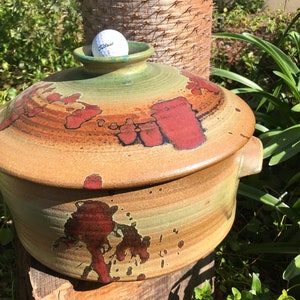 May include: A large, round, ceramic casserole dish with a lid. The dish has a brown, green, and red glaze with a speckled pattern. A white golf ball is sitting on top of the lid.