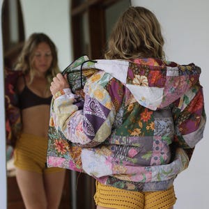 Puede incluir: Una chaqueta de patchwork colorida con capucha, con estampados florales y geométricos en tonos morados, verdes y rosas. La chaqueta se combina con shorts de crochet amarillos. La imagen se refleja en un espejo.