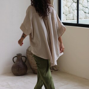 Puede incluir: Una camisa beige de gran tama&ntilde;o con mangas anchas y pantalones verde oliva. La camisa tiene una apariencia texturizada. La persona camina descalza sobre un suelo de color claro. Dos jarrones grandes de color marr&oacute;n oscuro est&aacute;n en el fondo.