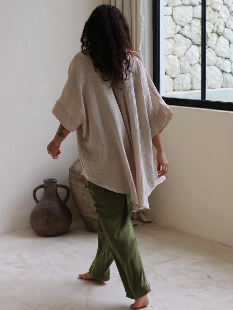 Puede incluir: Una camisa beige de gran tama&ntilde;o con mangas anchas y pantalones verde oliva. La camisa tiene una apariencia texturizada. La persona camina descalza sobre un suelo de color claro. Dos jarrones grandes de color marr&oacute;n oscuro est&aacute;n en el fondo.