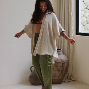 Puede incluir: Una mujer con una camisa beige abotonada de gran tama&ntilde;o, pantalones verde oliva y una parte superior negra. La camisa tiene un aspecto texturizado. Los pantalones son de color liso con un ligero brillo. La mujer sonr&iacute;e y tiene el pelo largo y oscuro.